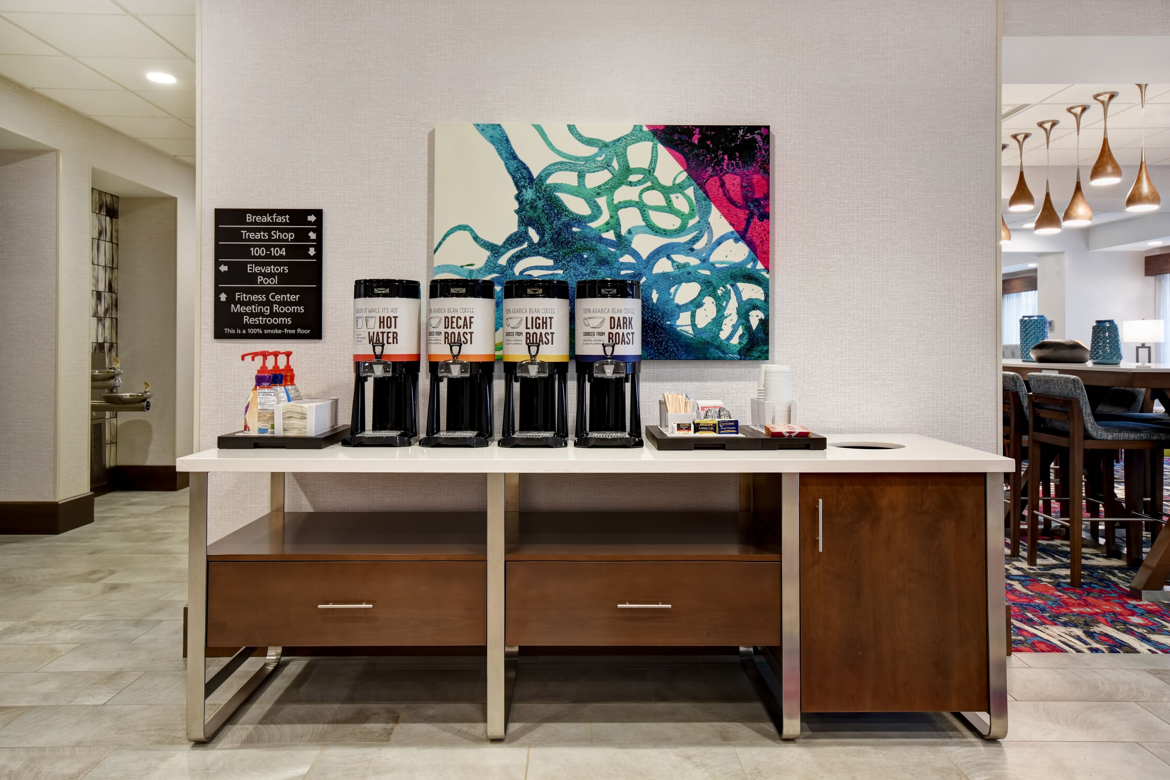 A hotel reception area with a coffee station featuring four coffee dispensers labeled 'Hot Water,' 'Decaf Roast,' 'Light Roast,' and 'Dark Roast.' There is a sign listing amenities such as breakfast, treats shop, elevators, pool, fitness center, meeting rooms, and restrooms.