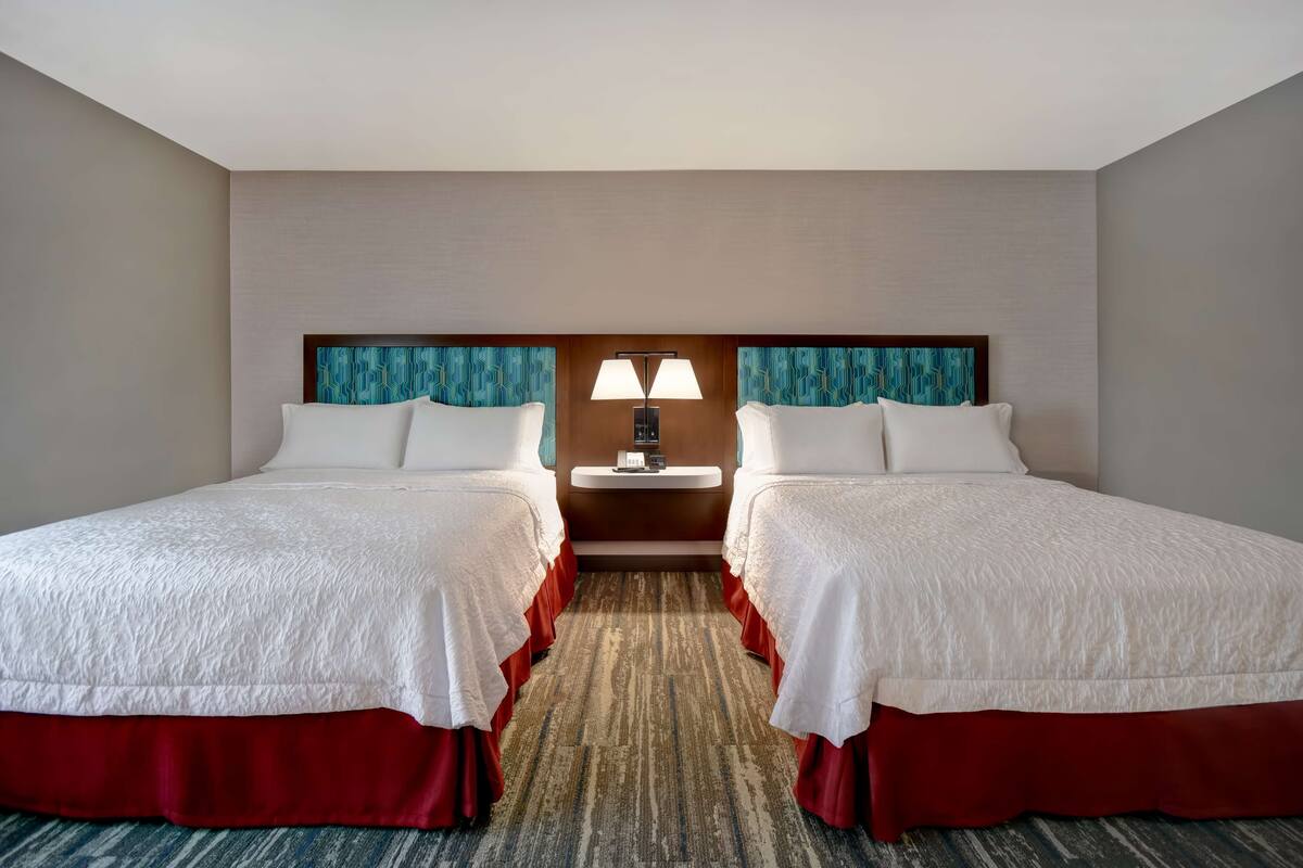 A hotel room with two beds, a nightstand with a lamp, and a carpeted floor.