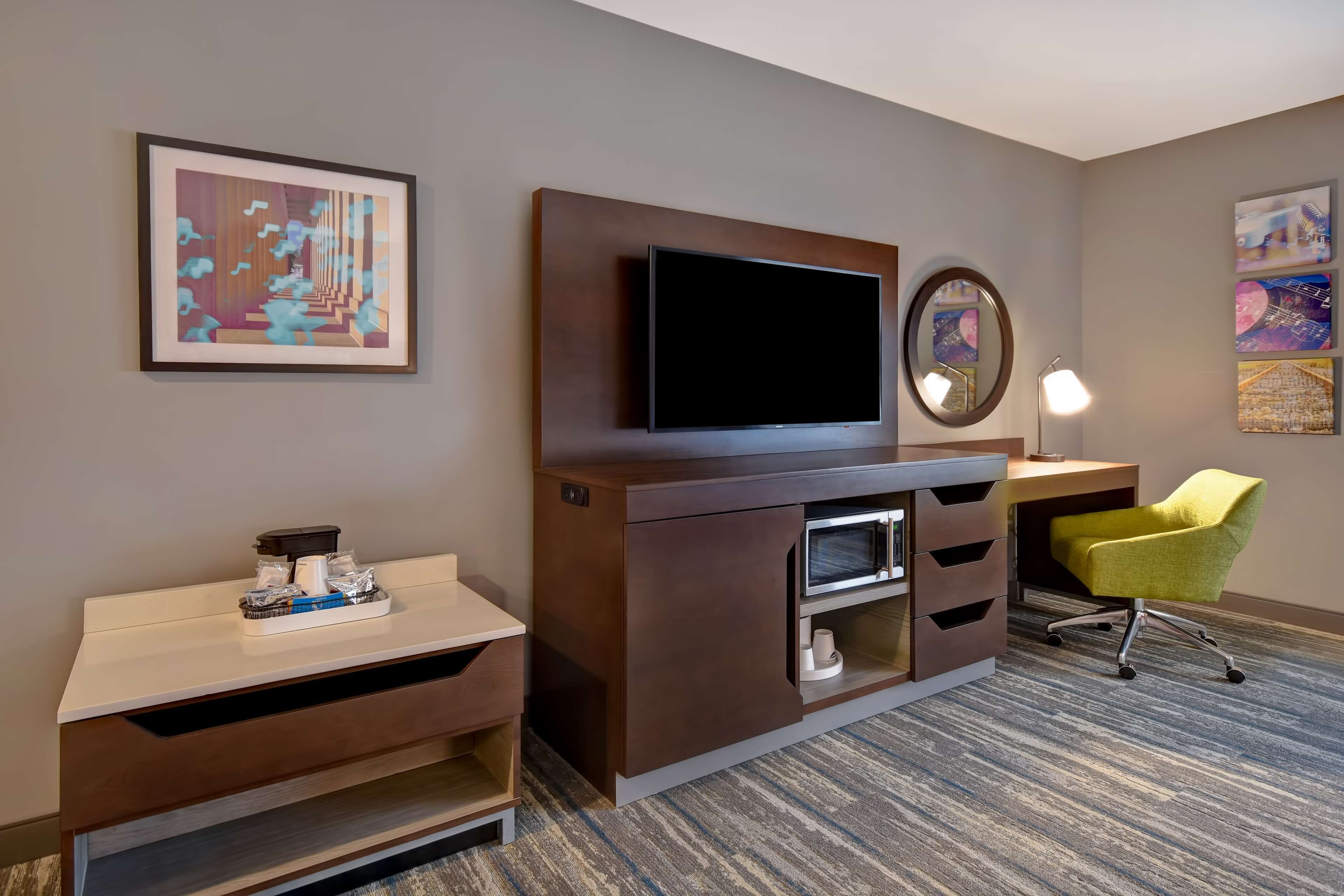 Habitación de hotel con televisión de pantalla plana montada en un mueble de madera, un escritorio con lámpara, una silla amarilla y un cuadro abstracto enmarcado en la pared.