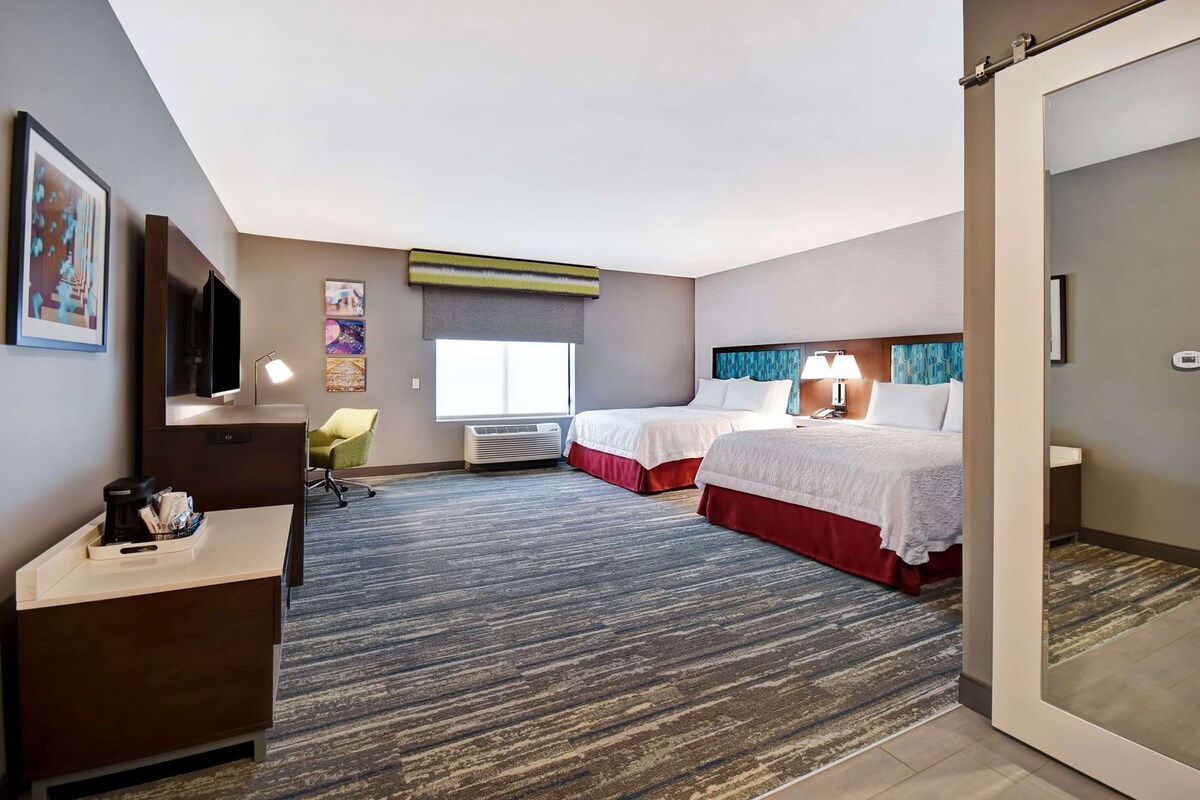 A hotel room with two beds, a desk with a chair, a TV, and a window with blinds.