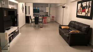 A modern living room with a black leather sofa, a TV on a stand, and a dining area with red stools. - Santa Maria del Mar, Terramar 315, 204 (Urb Santa Maria del Mar)