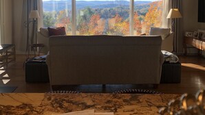 Uma sala de estar aconchegante com uma grande janela que oferece uma vista panorâmica de folhagem outonal e montanhas.