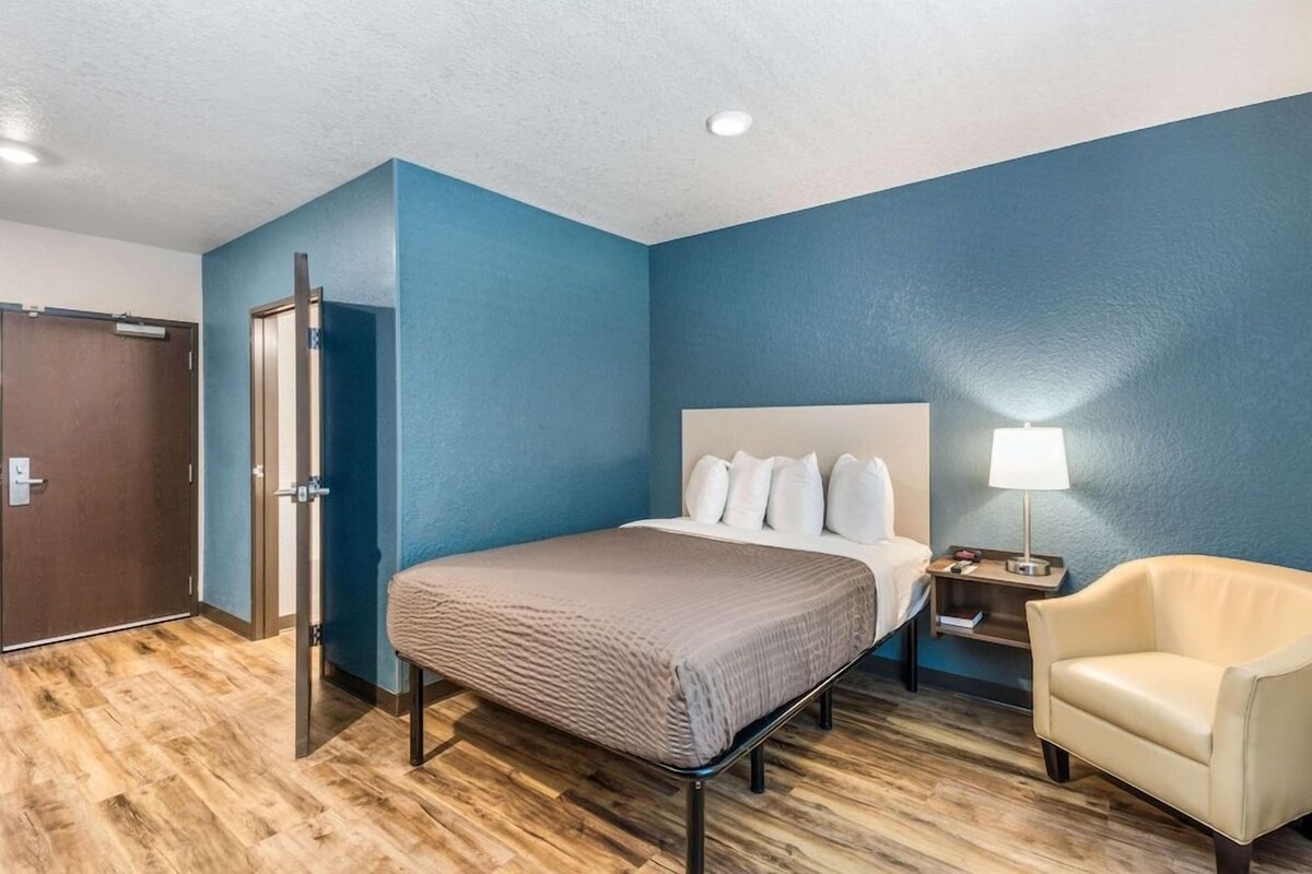 A hotel room with a bed, a chair, a nightstand, and a door.