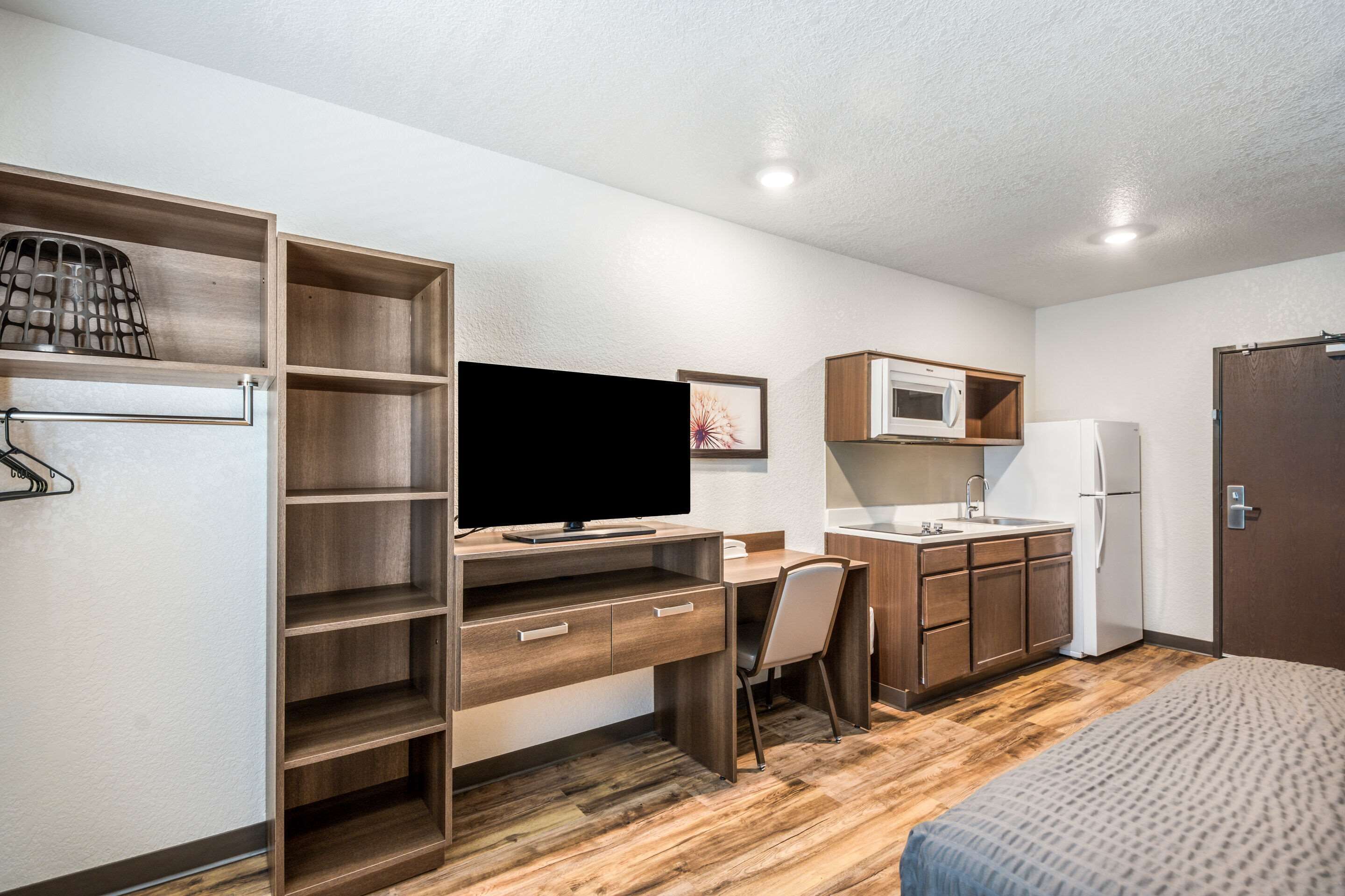 A compact room with a wooden desk, a television, a kitchenette with a microwave and refrigerator, and a bed.