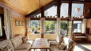A cozy cabin interior with wooden walls, a sofa, chairs, a table, and large windows offering a view of greenery. - Beautiful private villa for 6 people with WIFI, TV, balcony and pets allowed (Außertal)