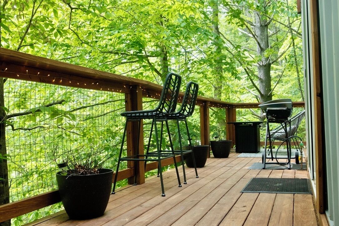 Una terraza de madera con sillas y una parrilla, rodeada de abundante vegetación.
