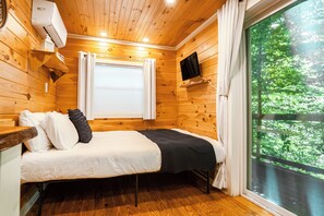 A wooden cabin room with a bed, a window with curtains, a mounted television, and a view of greenery outside.