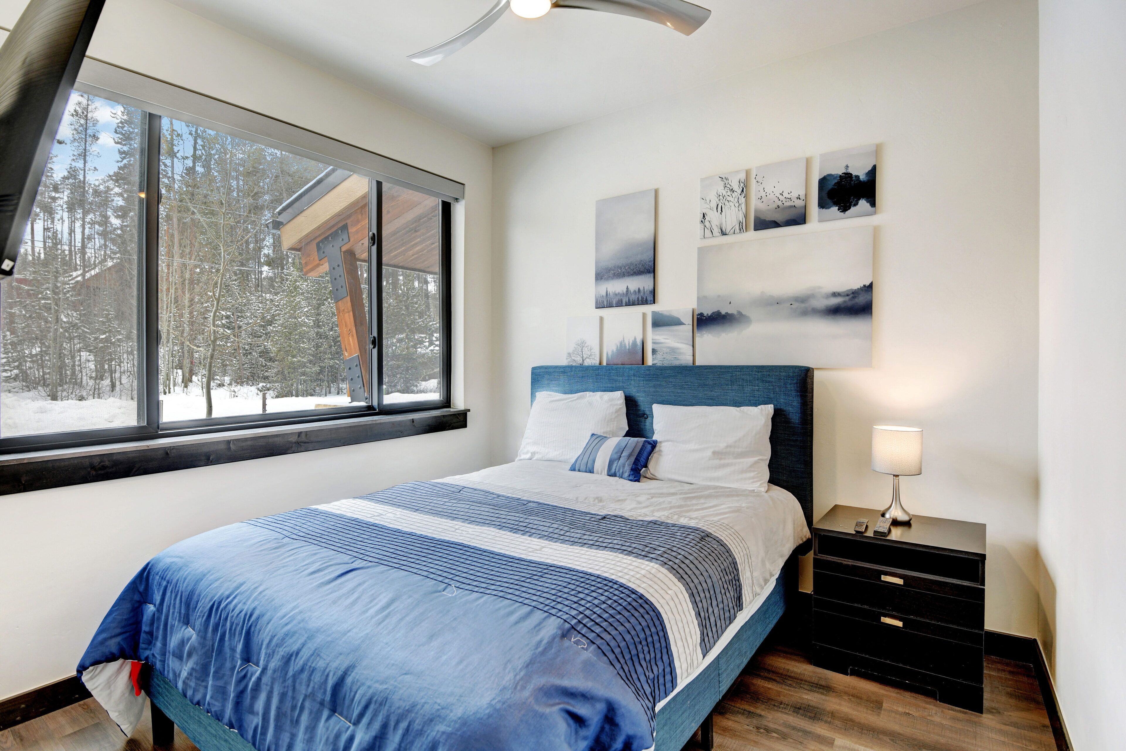A bedroom with a large window, a bed with a blue and white bedspread, a headboard, a nightstand with a lamp, and wall art.