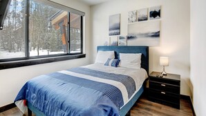 A bedroom with a large window, a bed with a blue and white bedspread, a headboard, a nightstand with a lamp, and wall art.