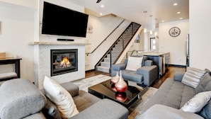 A modern living room with a fireplace, grey sofas, a flat-screen TV, and a staircase leading to another level.