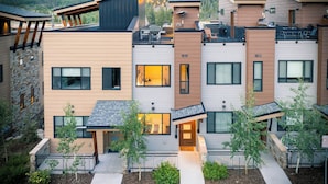 A modern multi-story residential building with a mix of wood and stone facades, large windows, and a rooftop deck with outdoor seating.