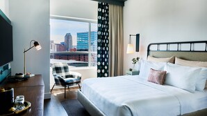 A hotel room with a large bed, a desk with a lamp, a chair, and a view of the city skyline.