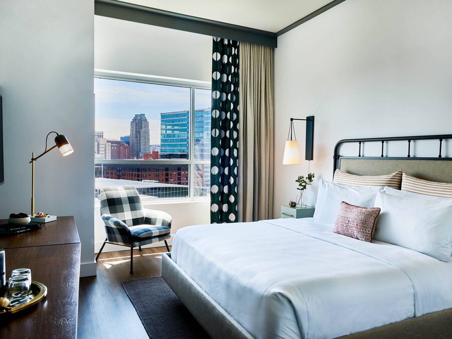 Et hotelværelse med en stor seng, et skrivebord med lampe, en stol og udsigt til byens skyline.