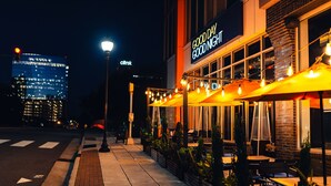 A nighttime view of a restaurant with outdoor seating, a visible street, and a city skyline in the background.