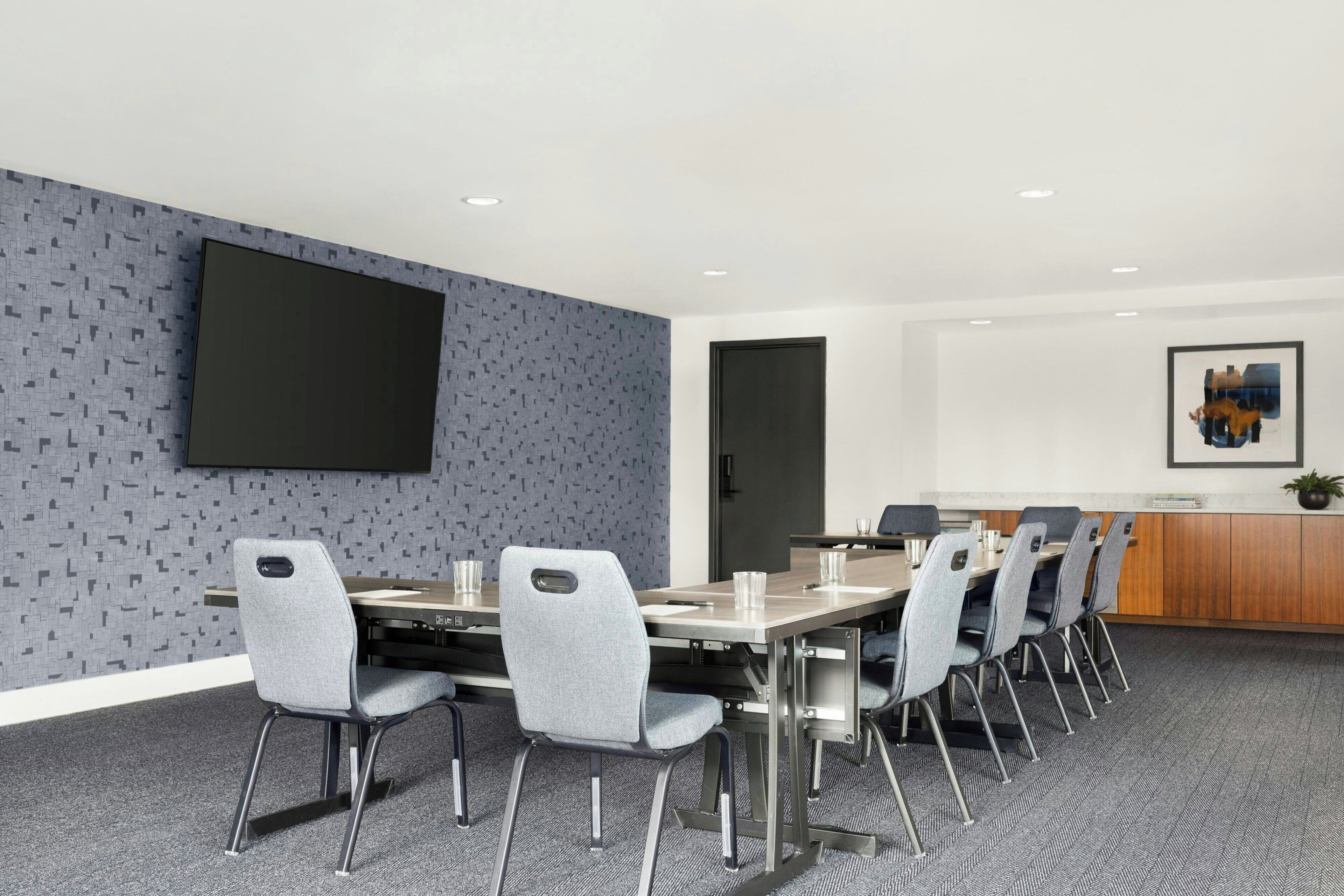 A conference room with a long table, grey chairs, a flat-screen TV, a wooden cabinet, and a patterned wall.