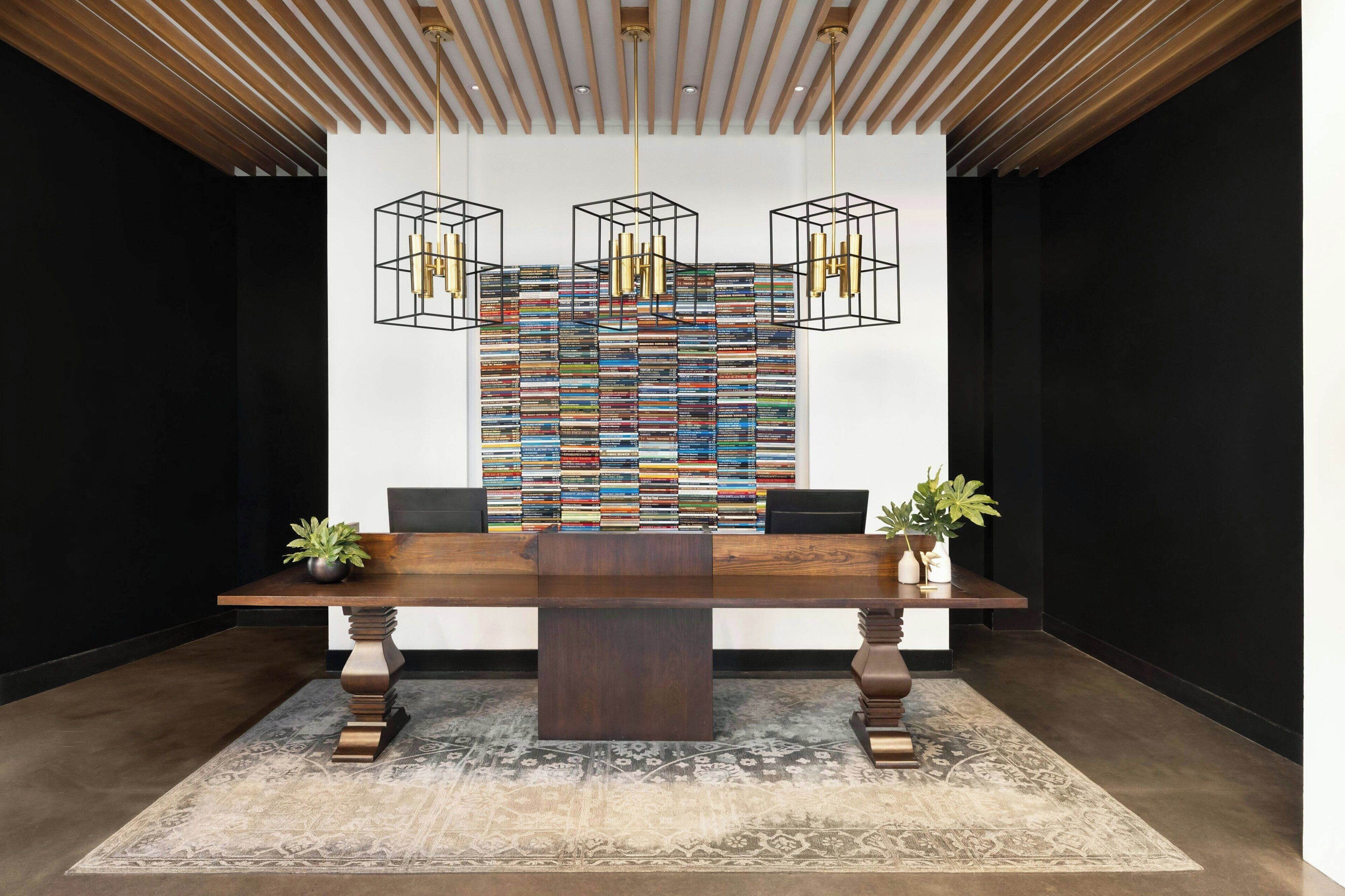A modern reception area with a wooden ceiling, a large bookshelf filled with books, two potted plants, and a dark wooden reception desk.