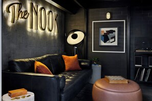 A modern lounge area with a leather sofa, a round ottoman, a chessboard, and a wall-mounted lamp.