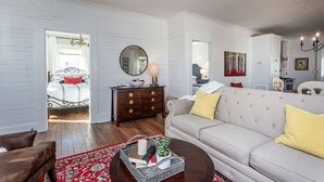 A living room with a beige sofa, a wooden coffee table, a red patterned rug, a dark wooden side table with a mirror, and a white wall with a painting. - Highland Gold+ Gather Family+Friends+make memories (Cleveland)