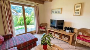 Una sala de estar con un sofá, un televisor, una mesa con velas, una planta y vista a espacios verdes al exterior.