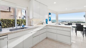 A modern kitchen with white cabinetry, a large window overlooking a scenic view, and a dining area with black chairs. - Ocean Views, Rooftop Deck, Fire Pit, Game Room, AC (Carlsbad)