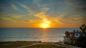 A sunset over the ocean with a view of a coastal area, including a building with a tiled roof and a pathway leading to the beach. - Ocean Views, Rooftop Deck, Fire Pit, Game Room, AC (Carlsbad)