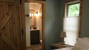 A bedroom with a bed, a nightstand, a lamp, a window with blinds, and a wooden door leading to a bathroom. - Your search ends with KSW Cabin. All a group needs, incl. 5 bathrooms. Sleeps 14 (Campton)