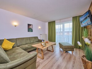 Un salon moderne avec un canapé vert, une table basse en bois, un petit pouf, un téléviseur fixé au mur et une grande fenêtre avec des rideaux.