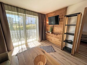 Una sala moderna con suelo de madera, un televisor montado en la pared, una mesa de centro de madera y un ventanal corredero que da a un espacio exterior.