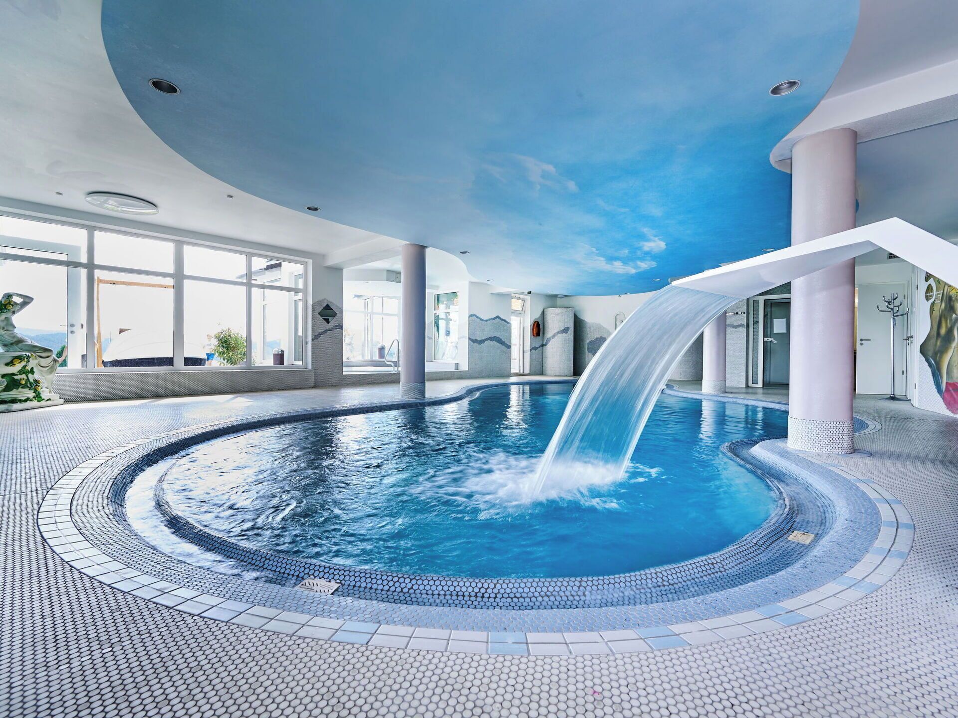 Una gran piscina interior amb cascada, envoltada per un paviment de rajoles i grans finestres.
