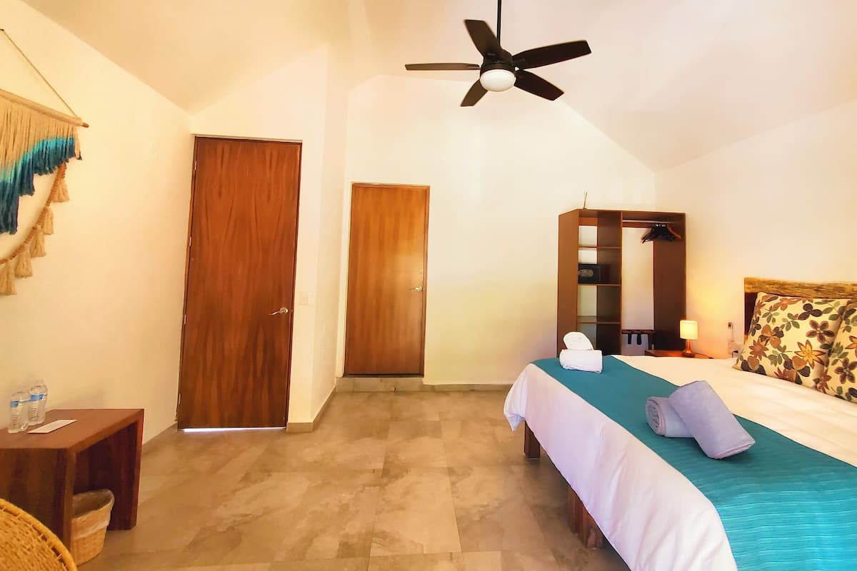 Une chambre d’hôtel avec un lit, un ventilateur de plafond, une porte en bois, une petite table avec un panier et une décoration murale.