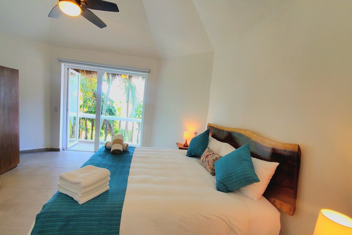 Une chambre spacieuse avec un grand lit, un ventilateur de plafond et une vue sur la verdure par la fenêtre.
