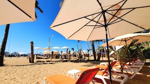 Une plage bordée de rangées de transats et de parasols, de palmiers, et un ciel d’un bleu limpide.