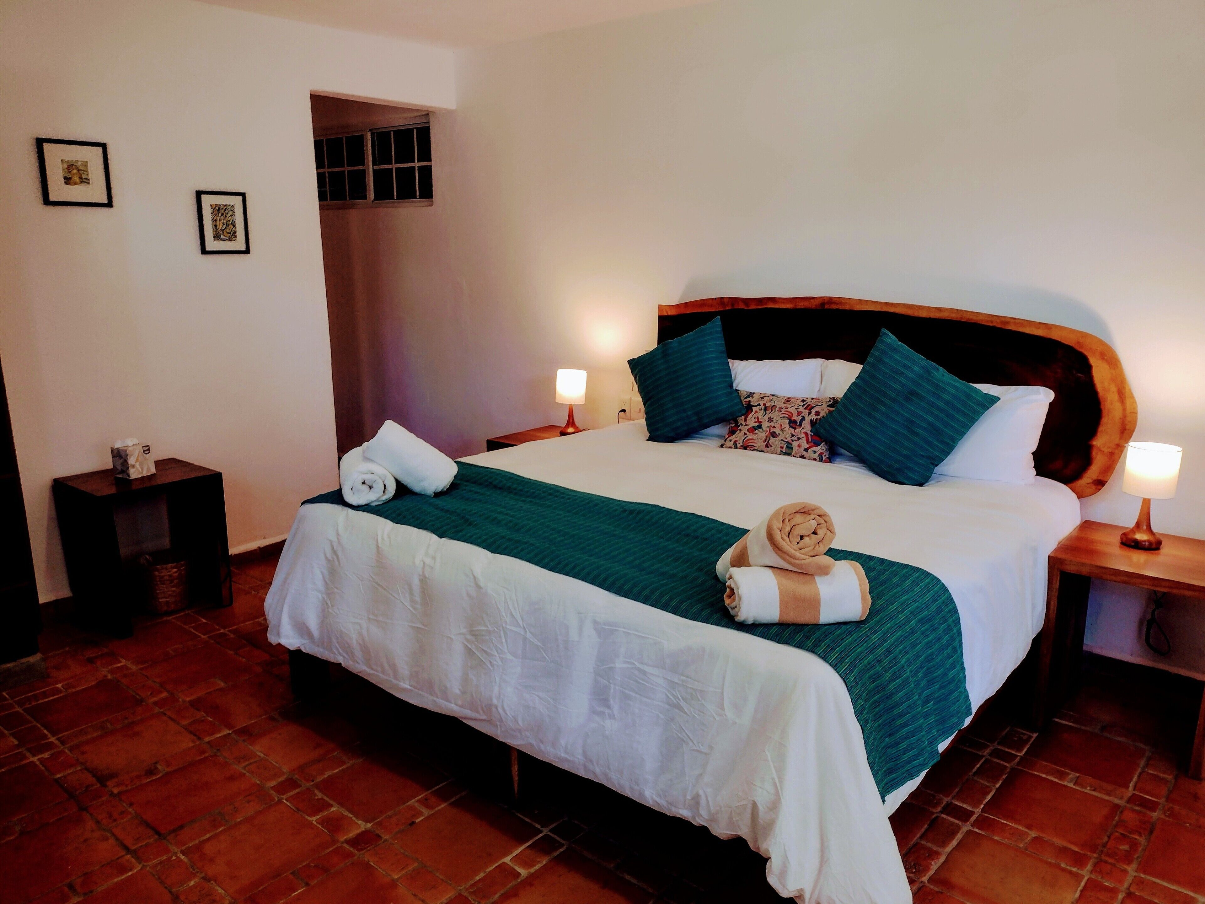 Una cama bien tendida con una colcha verde y blanca, dos almohadas y dos toallas enrolladas. Hay dos lámparas de noche, una mesita con una caja de pañuelos y un marco de fotos en la pared.
