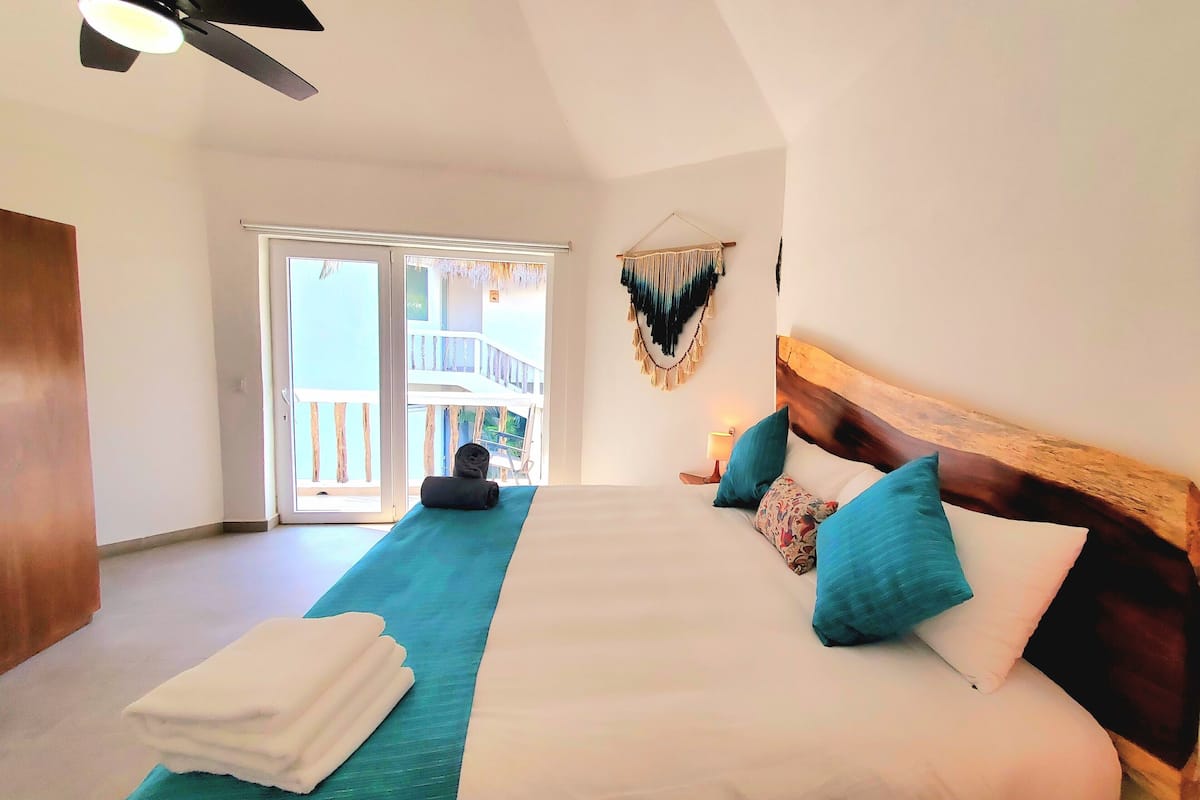 Une chambre d’hôtel avec un grand lit, un ventilateur de plafond, un balcon avec une balustrade et un tableau accroché au mur.