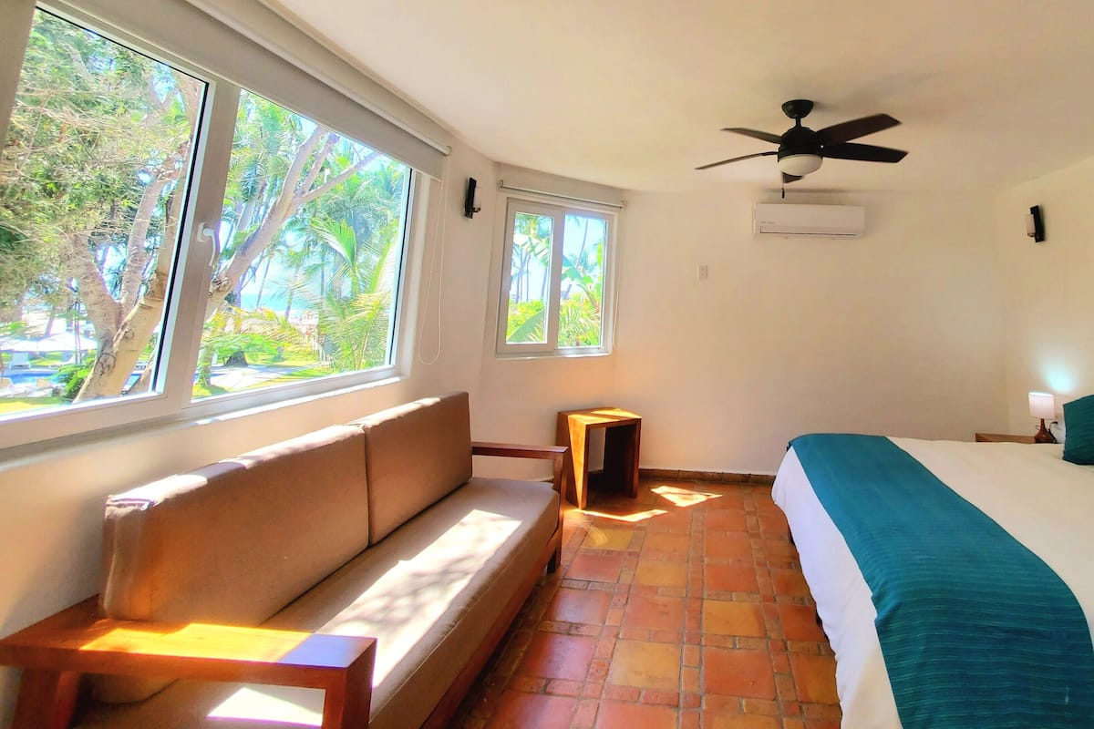 Une chambre à coucher avec un lit, un banc, un ventilateur de plafond et de grandes fenêtres.