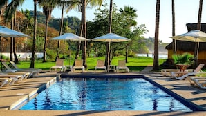 Une piscine rectangulaire bordée de chaises longues et d’ombrelles, entourée de palmiers et offrant une vue sur une étendue d’eau.