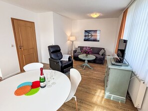 A living room with a sofa, a black chair, a small round table with a vase, a TV on a stand, and a radiator.