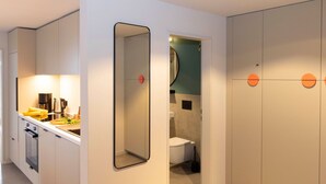 Eine moderne Küche mit Einbauofen, Kaffeemaschine und Spüle. An der Wand hängt ein Spiegel und eine Tür führt zu einem Badezimmer mit Toilette und Dusche.