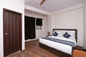 Une chambre d’hôtel avec un plancher en bois, un lit avec une literie blanche et des oreillers bleus, une table de chevet avec une lampe et une fenêtre avec des rideaux noirs.