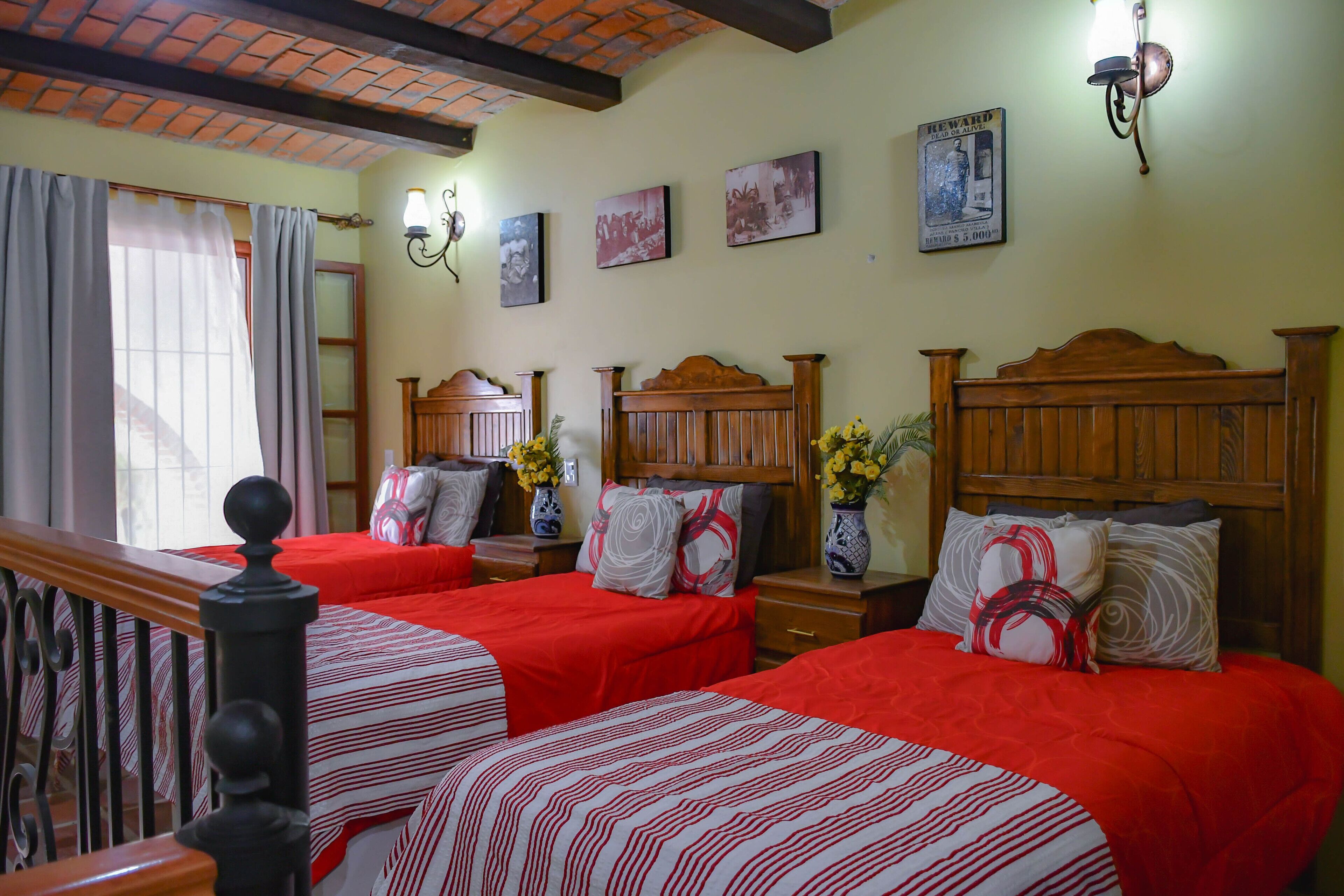 A hotel room with two beds, each with a wooden headboard, red bedding, and decorative pillows. There are wall-mounted light fixtures and framed pictures on the wall.