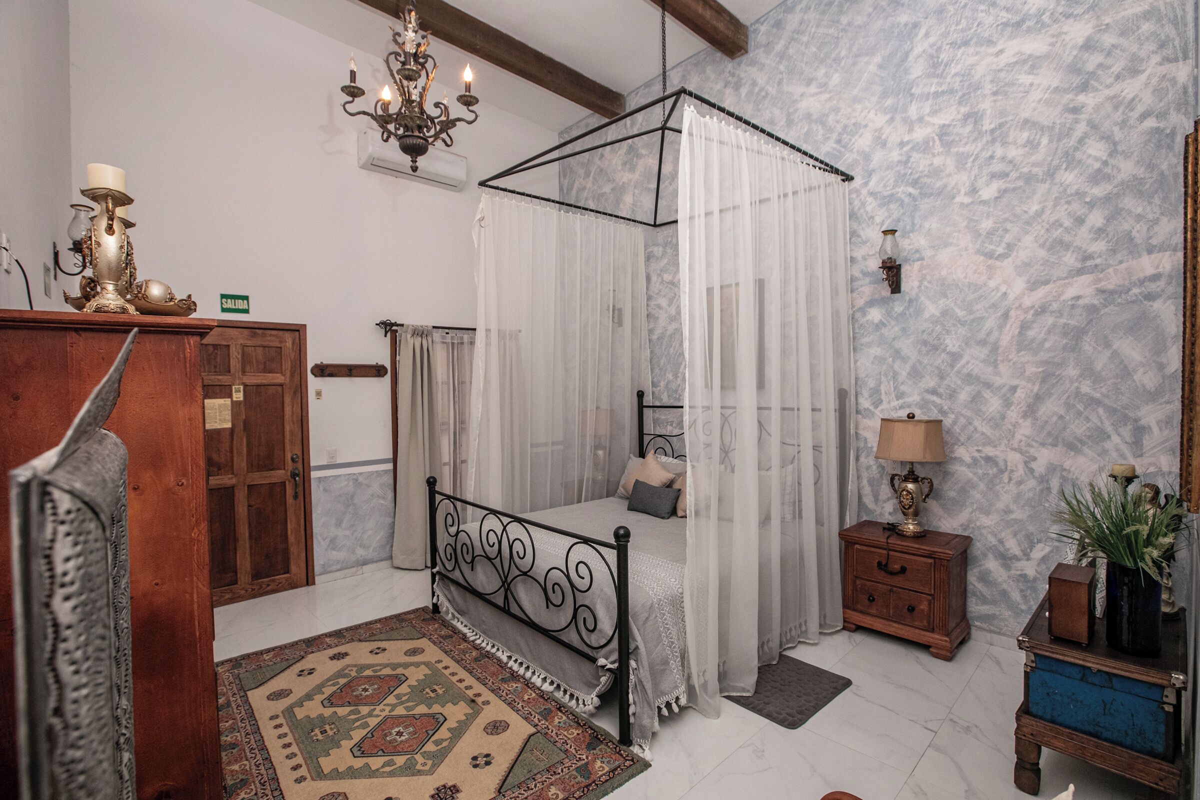 A bedroom with a four-poster bed, a wooden dresser, a nightstand, a chandelier, and a marble-patterned wall.