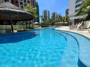 Una piscina con borde de azulejos, rodeada de sillones de descanso y palmeras.