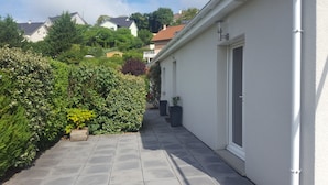 Un étroit sentier mène à un bâtiment blanc doté d’une porte et d’une fenêtre, le tout étant entouré d’une végétation luxuriante et de plantes en pot.