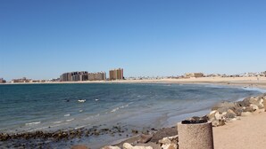 A coastal view with a sandy beach, rocky shoreline, and buildings in the background. - Marina Pinacate 306 Sandy Cove Refuge Wonderful Ocean View Condo (Puerto Peñasco)