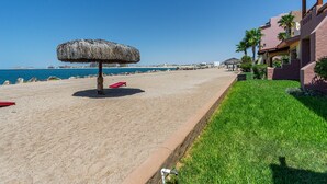 Une plage avec un parasol en chaume, une chaise longue rouge et un bâtiment doté d’un jardin de palmiers.