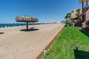 A beach with a thatched umbrella, a red lounge chair, and a building with a palm tree garden.