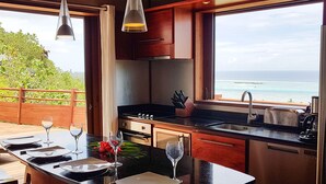 A modern kitchen with a view of the ocean, featuring a central island, stainless steel appliances, and wooden cabinetry. - Villa Tea by Tahiti Homes (Moorea-Maiao)