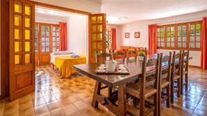 Un coin repas avec une table et des chaises en bois, un lit recouvert d’une couverture jaune et une fenêtre ornée de rideaux orange.
