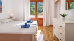 Une chambre d’hôtel avec deux lits, un plancher en bois, une commode blanche, une plante en pot et une grande fenêtre avec des rideaux.