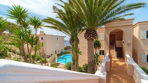 Vista de la piscina con una palmera, un edificio con balcón y cielo despejado.